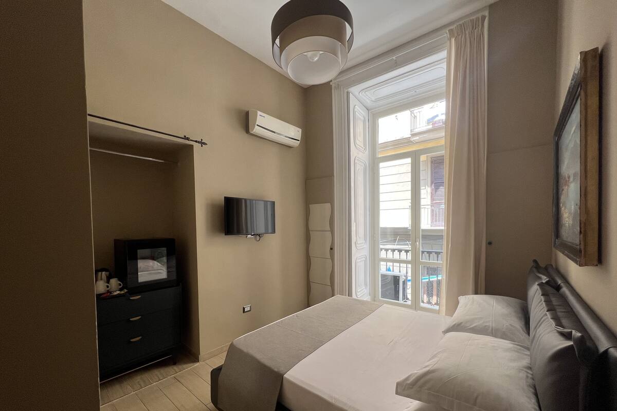 A hotel room with a bed, a TV, an air conditioner, a wardrobe, and a window with a view of buildings.