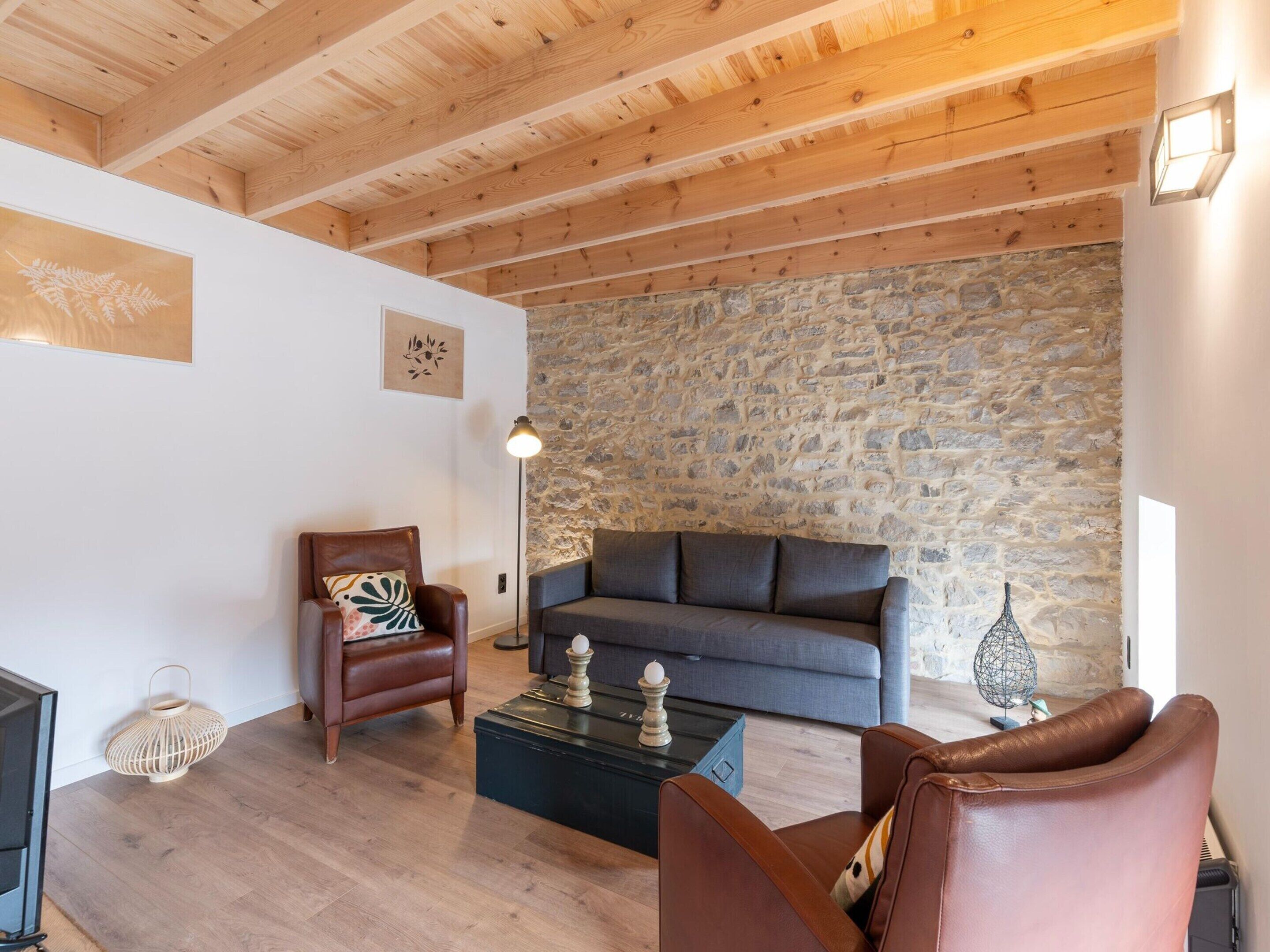 A cozy living room with a stone wall, wooden ceiling beams, a leather armchair, a grey sofa, and a coffee table.