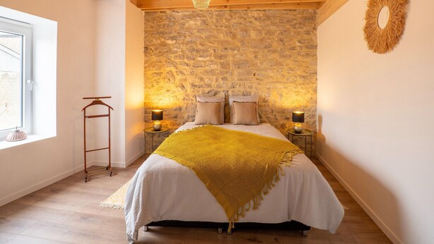 A bedroom with a stone wall, wooden ceiling beams, a bed with a yellow blanket, two bedside lamps, and a hanging light fixture. - Spacious Holiday Home in Doische With Private Pool (Doische)