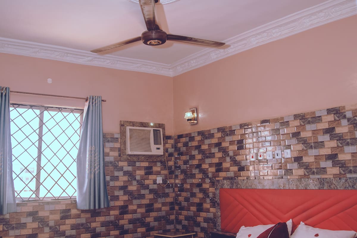 A bedroom with a bed, a ceiling fan, an air conditioning unit, a window with curtains, and a tiled wall.