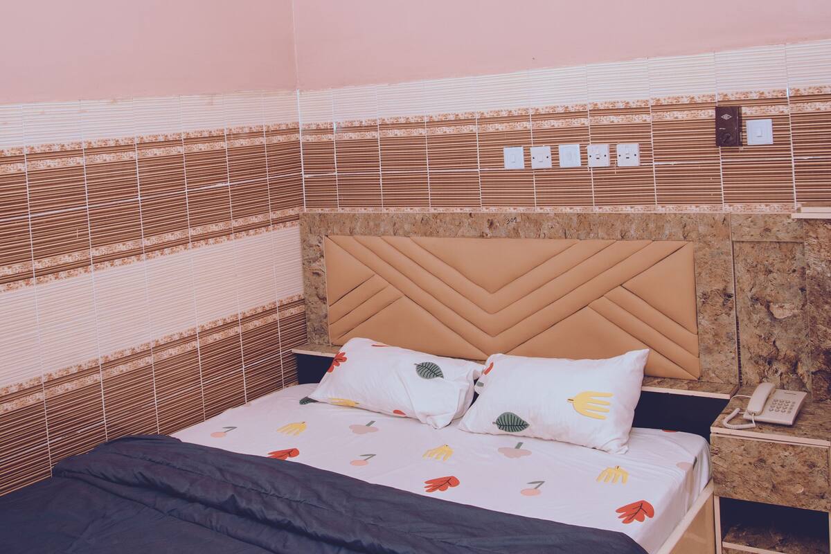 A bed with a patterned headboard, a bedside table with a phone, and a wall with a decorative panel.