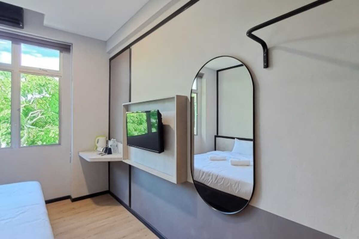 A bedroom with a bed, a mounted TV, a bedside table, a mirror, and a window with a view of greenery.