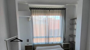 A hotel room with a bed, a ceiling fan, a window with sheer curtains, a radiator, a towel on the bed, a coat rack, and a shelf with toiletries. - Pensión Benavente. 4 - (Playa 1,5km / AP7-3min) (Moncofa)