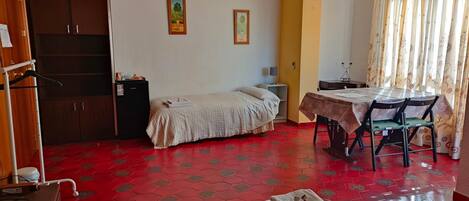 A room with a bed, a small table with a chair, a wardrobe, and a red tiled floor.