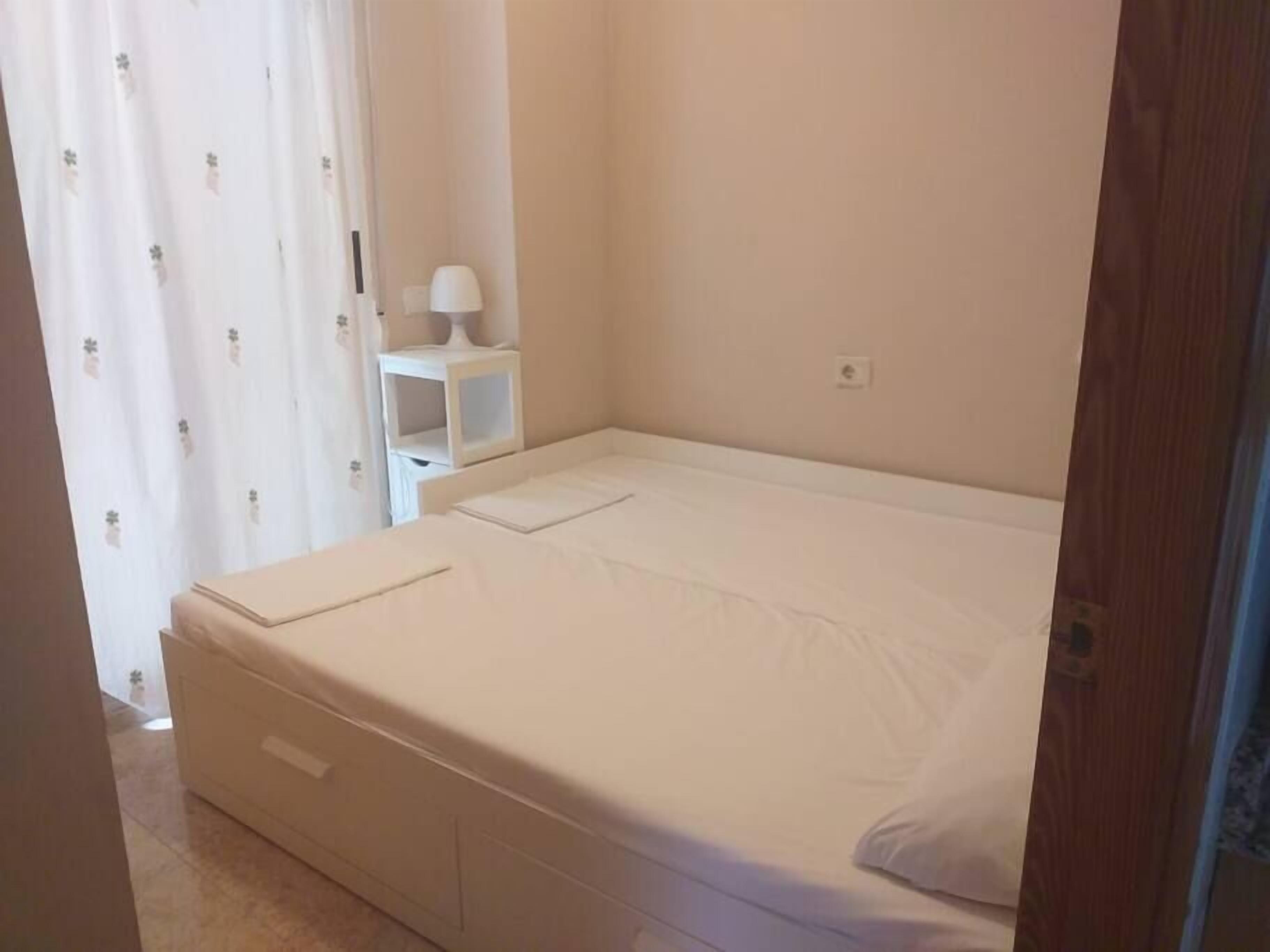 Un petit lit bien fait, avec un matelas et des oreillers blancs, une table de chevet avec une lampe et une petite étagère.