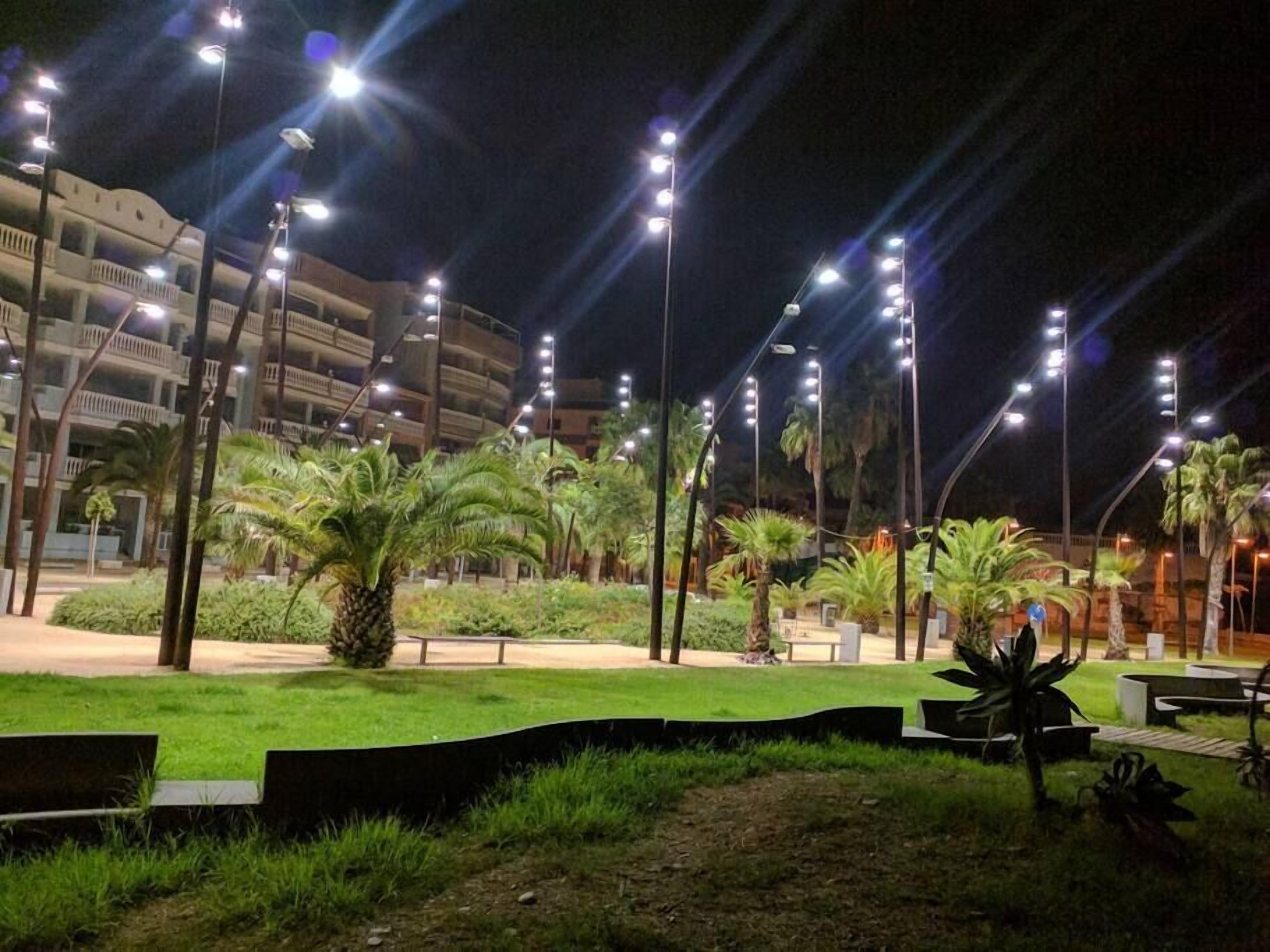 Un espacio exterior bien iluminado con palmeras, bancos y un edificio con varios balcones al fondo.