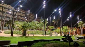 Un espacio exterior bien iluminado con palmeras, bancos y un edificio con varios balcones al fondo.