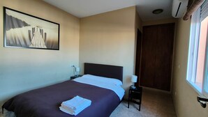 Habitación de hotel con cama, mesita de noche, lámpara, un cuadro en la pared y ventana con persianas.