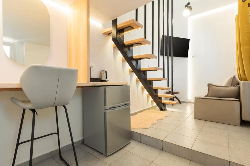 Un espacio habitable moderno con cocina, una silla de comedor, un refrigerador pequeño, un sofá y una escalera con escalones de madera y barandilla negra.