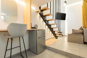 A modern living space with a kitchen area, a dining chair, a small refrigerator, a sofa, and a staircase with wooden steps and a black railing. - Mykonos Eight (Mykonos)