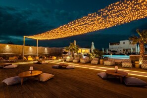 An outdoor seating area with round tables and cushioned chairs, a pool, and a string of lights against a dark sky. - Mykonos Eight (Mykonos)