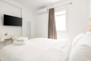 A hotel room with a bed, a flat-screen TV mounted on the wall, a bedside table with a lamp, and a window with curtains. - Mykonos Eight (Mykonos)