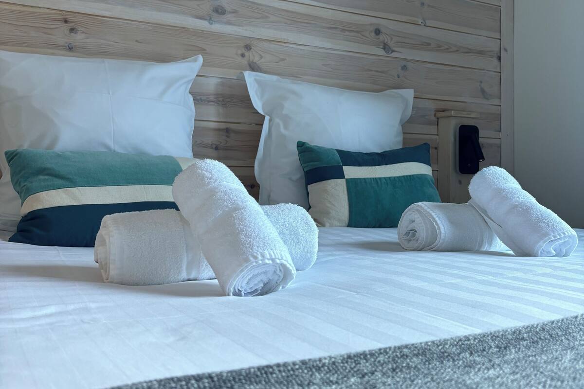 A neatly made bed with white linens, two pillows with a green and blue stripe, and two rolled white towels.