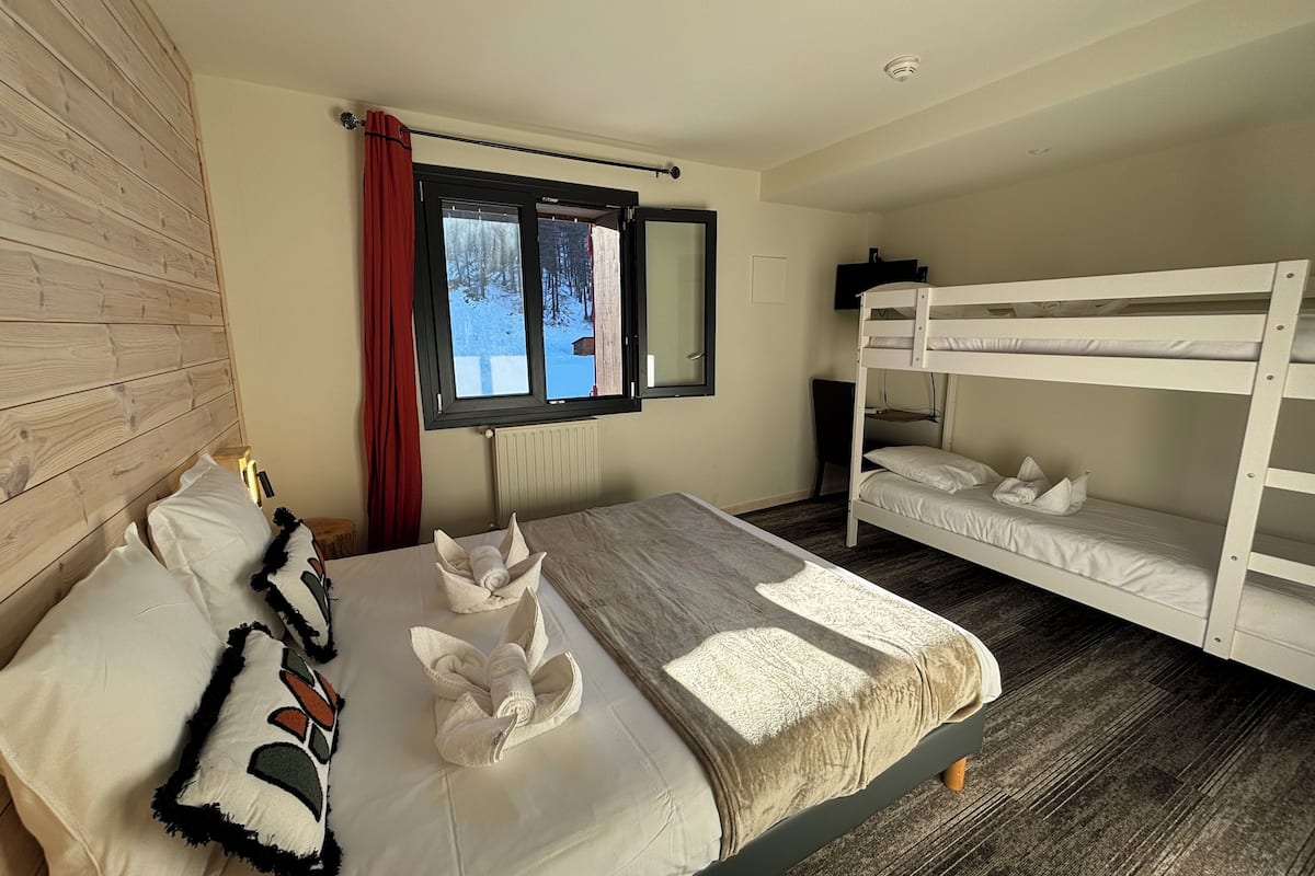 A hotel room with a wooden headboard, a large bed with white bedding, a window with a view of trees, a bunk bed, and a radiator.