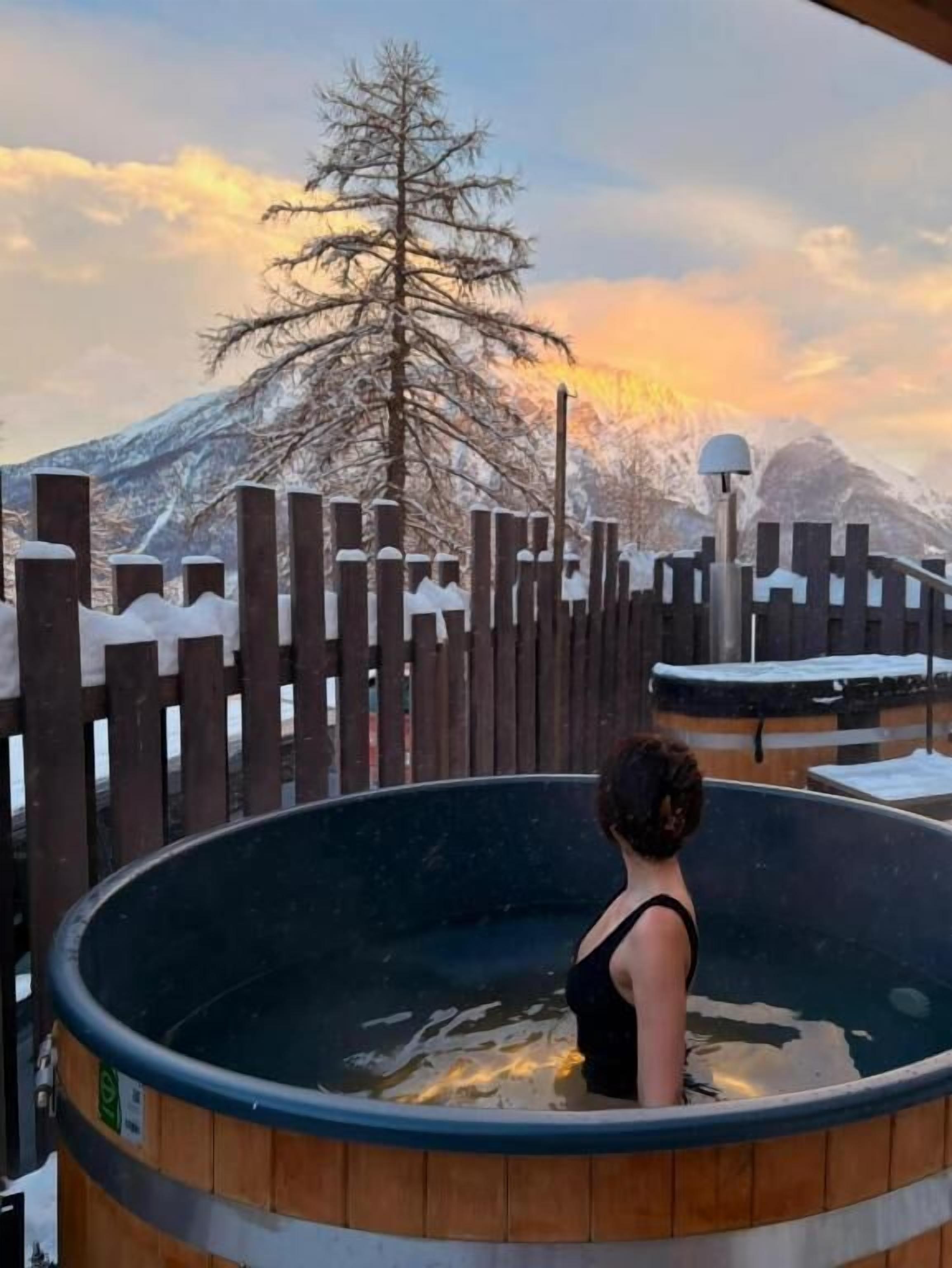 A person in a wooden hot tub with a view of a snow-covered mountain and a tree in the background. - Chalet Sainte Anne (La Condamine-Chatelard)