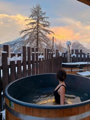 A person in a wooden hot tub with a view of a snow-covered mountain and a tree in the background. - Chalet Sainte Anne (La Condamine-Chatelard)