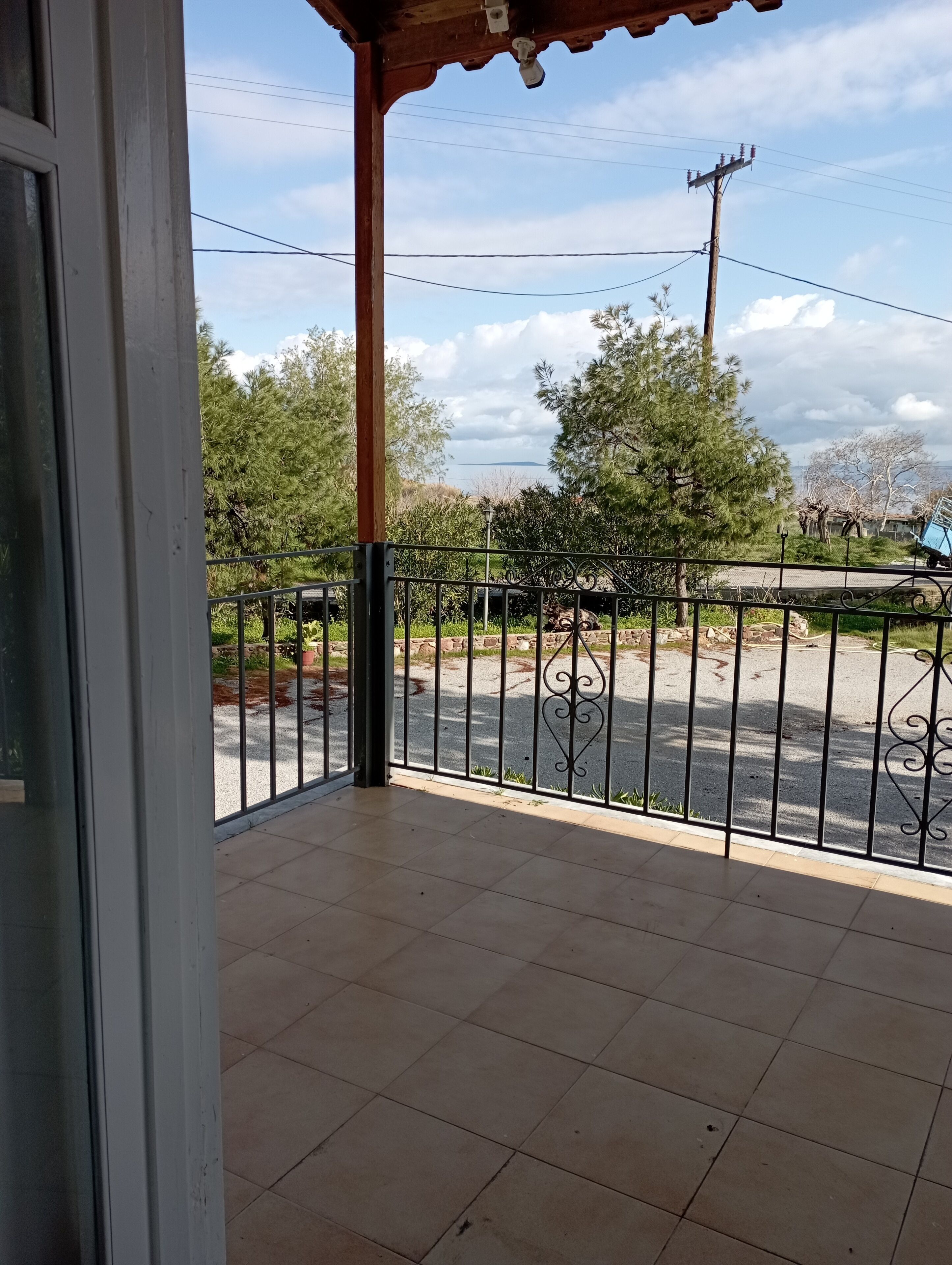 Ein überdachter Balkon mit gefliestem Boden, einem Metallgeländer und Blick auf einen Parkplatz mit Bäumen und bewölktem Himmel.