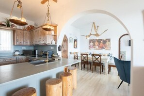 A modern kitchen with wooden cabinets, a central island, and a dining area with wooden chairs. - Blue Moon Mykonos Homes (Mykonos)