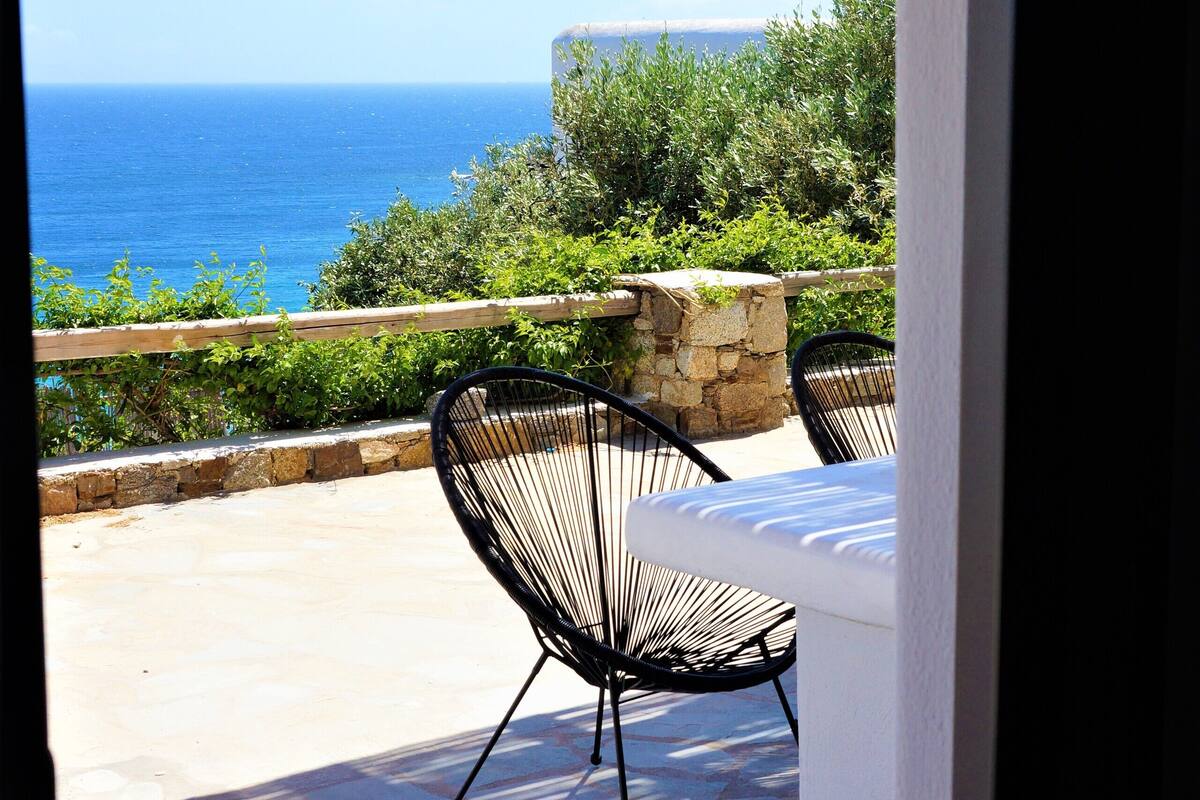 An open patio with a stone floor, a wooden pergola, a table with a white cushion, and two black chairs.
