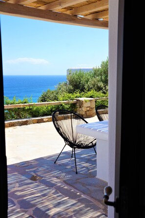 An open patio with a stone floor, a wooden pergola, a table with a white cushion, and two black chairs.