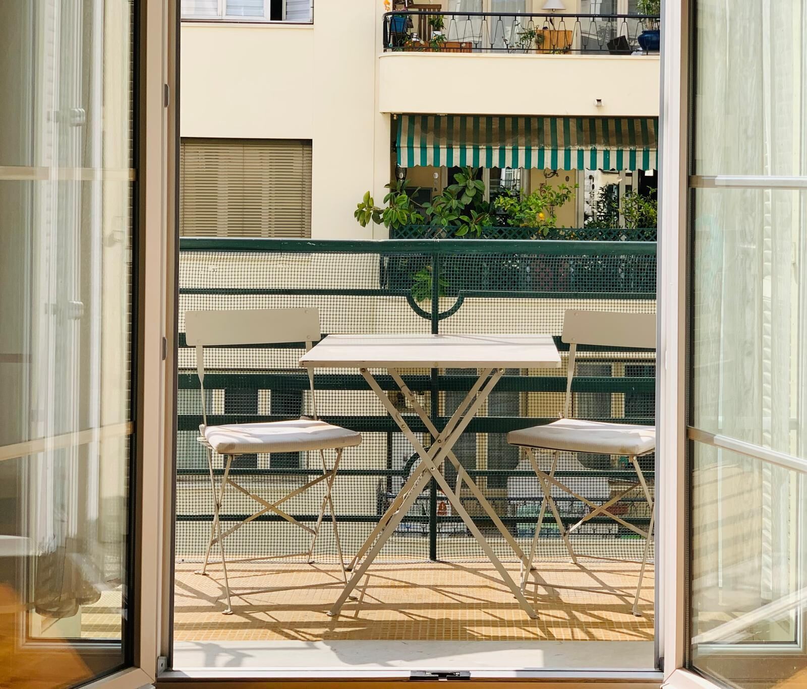 En balkon med et bord og to stole, et grønt sikkerhedsnet og udsigt til en bygning med markise.
