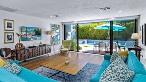 En moderne stue med stor sofa, sofabord og fjernsyn. Der er en balkon med pool og udendørs møbler.