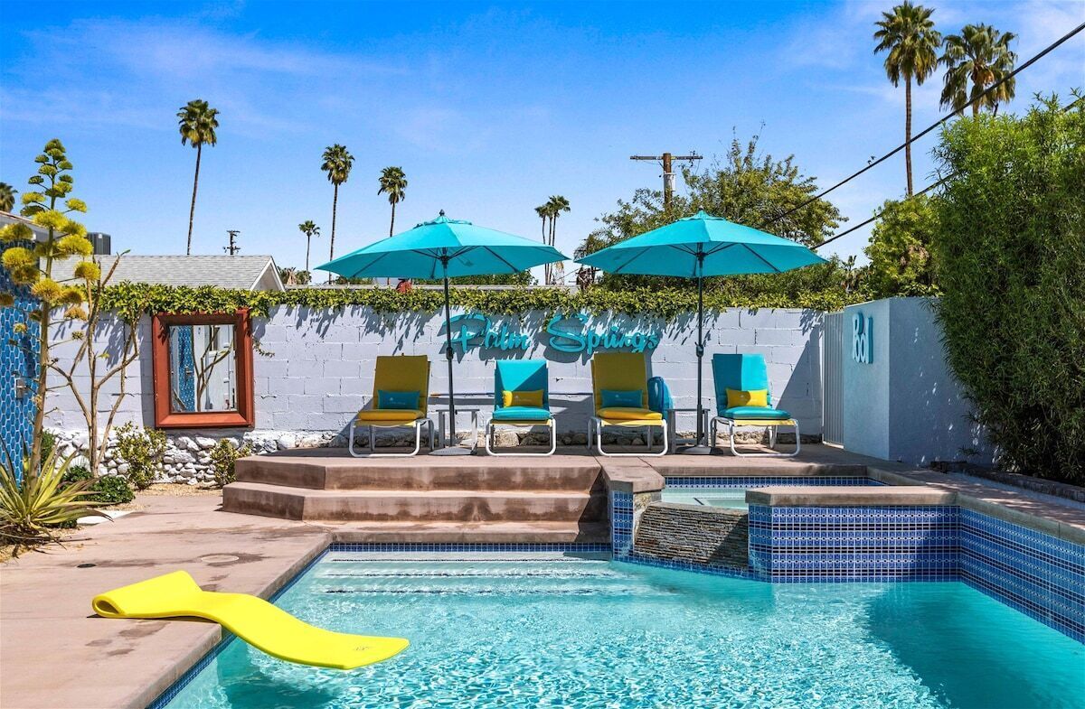 Zona de piscina con sillones de descanso y sombrillas, una piscina con azulejos y una pared con el texto "Palm Springs".