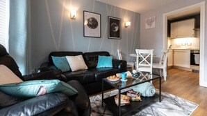 A modern living room with a black leather sofa, a glass coffee table, and a kitchen area visible through an open doorway.