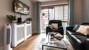 A living room with a black leather sofa, a glass coffee table, and a black armchair. There is a television mounted on the wall and a window with curtains.