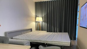 A compact hotel room with a bed, a small table, a lamp, and a view of the outside through a window with curtains. - Superbe Appartement Cosy - Plage des Minimes (La Rochelle)