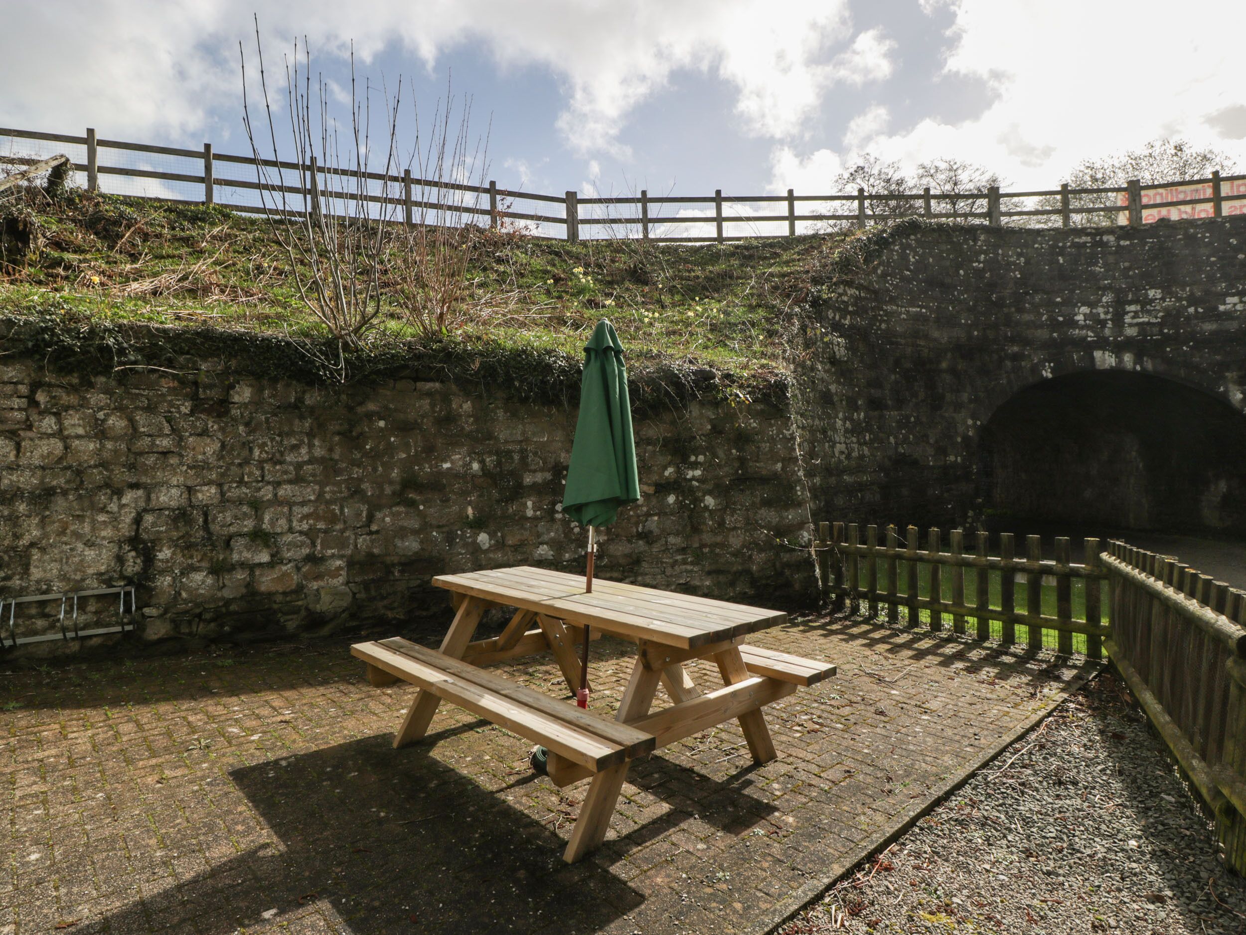 A picnic table with a green umbrella, set against a stone wall with a wooden fence and a brick pathway.