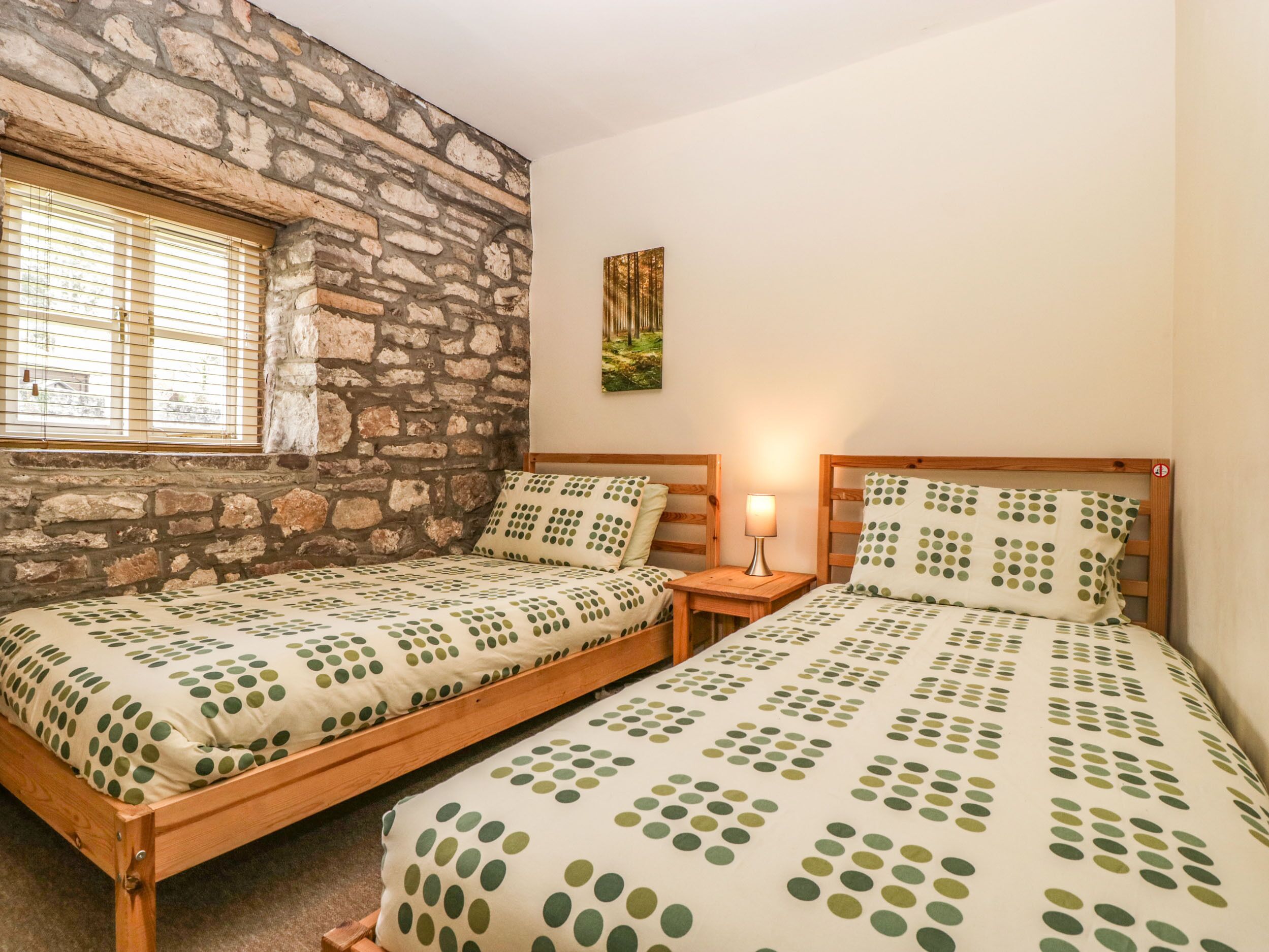 A bedroom with two wooden beds, a stone wall, a window with blinds, and a nightstand with a lamp.
