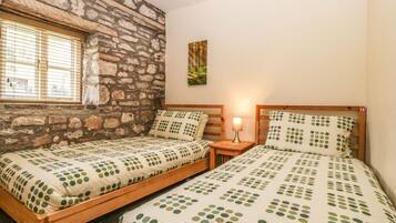 A bedroom with two wooden beds, a stone wall, a window with blinds, and a nightstand with a lamp.