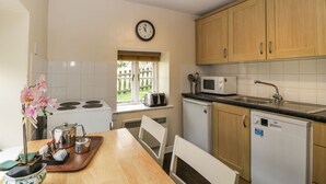 A kitchen with wooden cabinets, a microwave, a sink, a clock, and a window with a view of greenery. - Aqueduct Cottage (Pontypool)