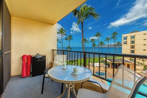 Ein Balkon mit einem Glastisch, zwei Stühlen, einem roten Koffer und einem schwarzen Lautsprecher. Im Hintergrund sind Palmen und ein Gebäude über dem Wasser zu sehen.