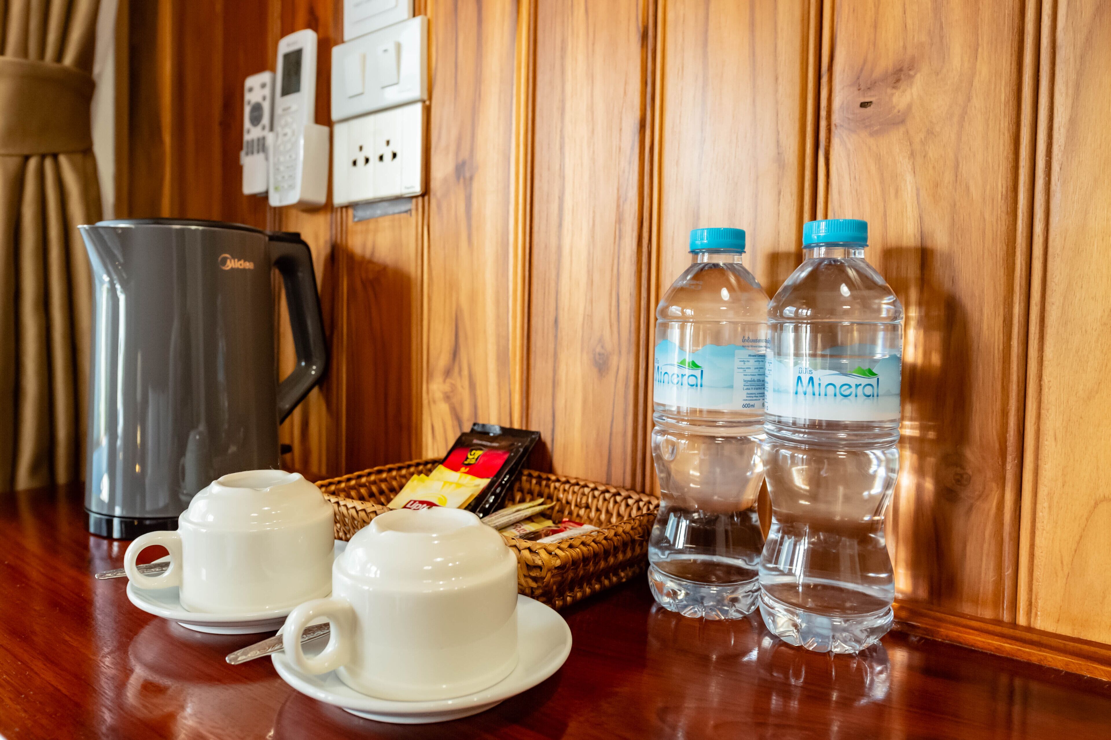 A wooden-paneled wall with electrical outlets, a kettle, two cups, a basket with a card, and two mineral water bottles.