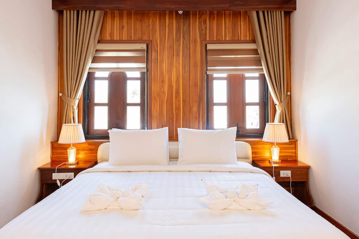 Une chambre d’hôtel avec une tête de lit en bois, deux lampes de chevet, un lit avec du linge de maison blanc et une fenêtre avec des rideaux.