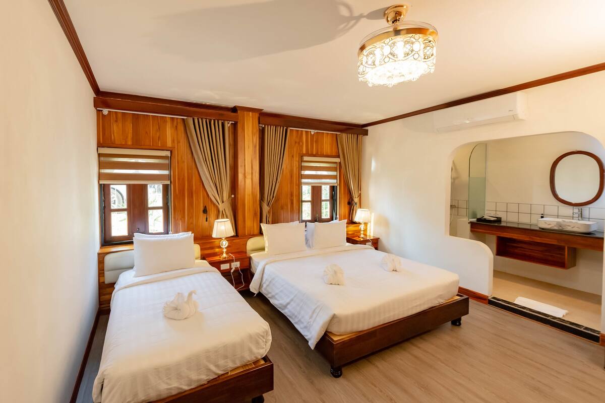 Une chambre d’hôtel avec deux lits, des boiseries, un lustre, et une salle de bain avec un miroir et un lavabo.