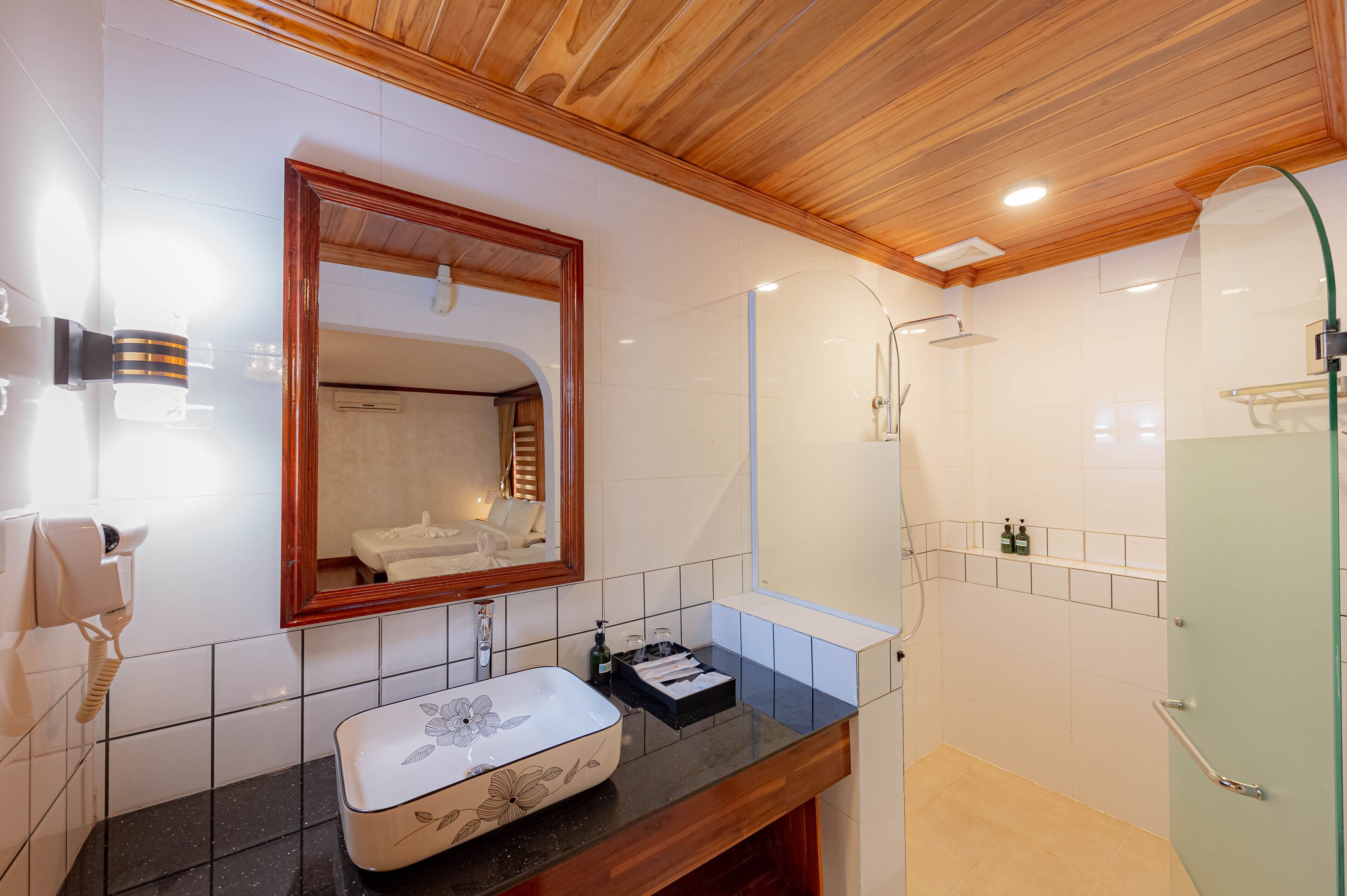 A bathroom with a wooden ceiling, a large mirror, a white sink with a floral design, a black countertop, and a glass shower enclosure.