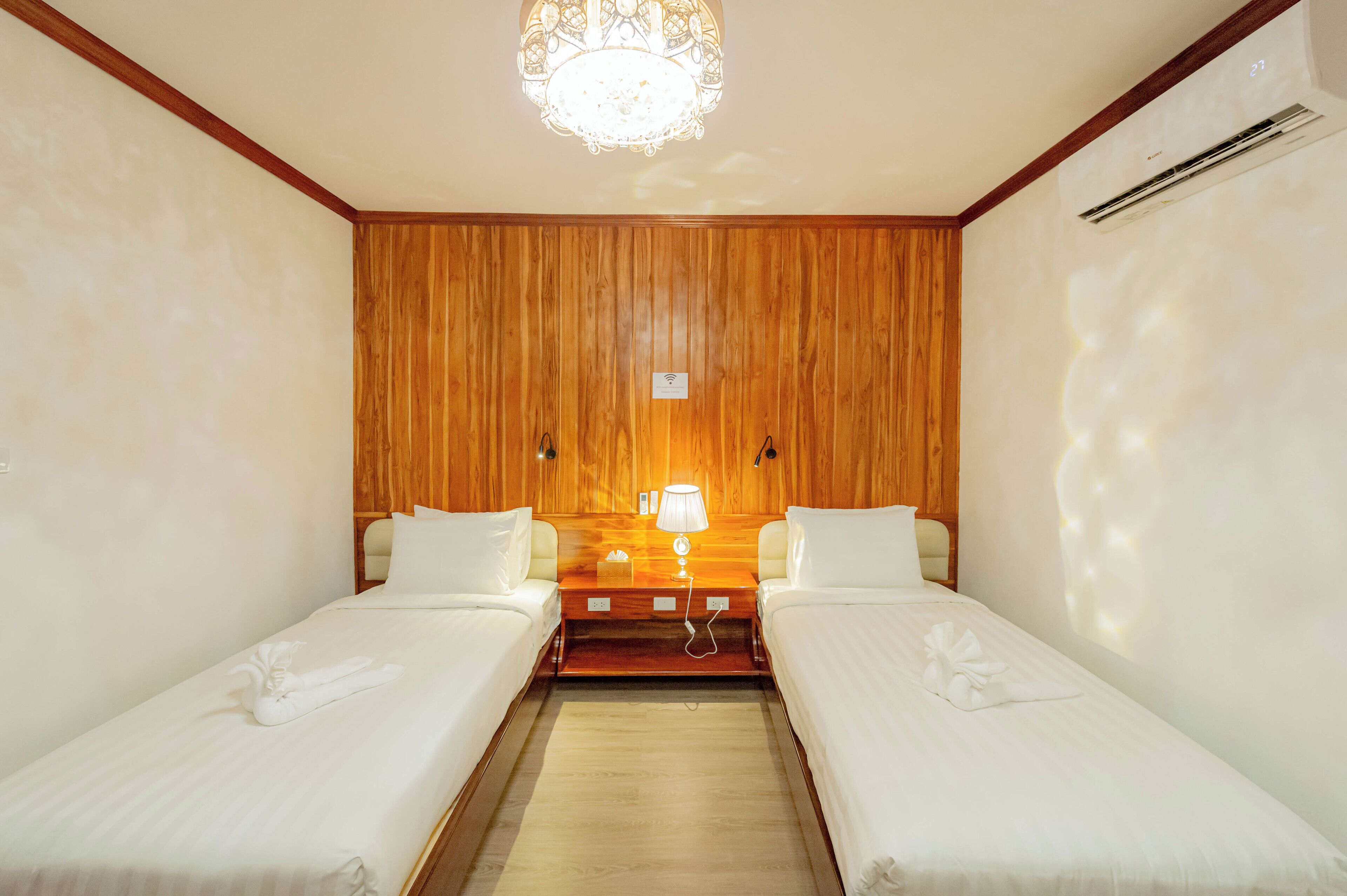 A hotel room with two single beds, wooden paneling on the walls, a chandelier, and a bedside table with a lamp.