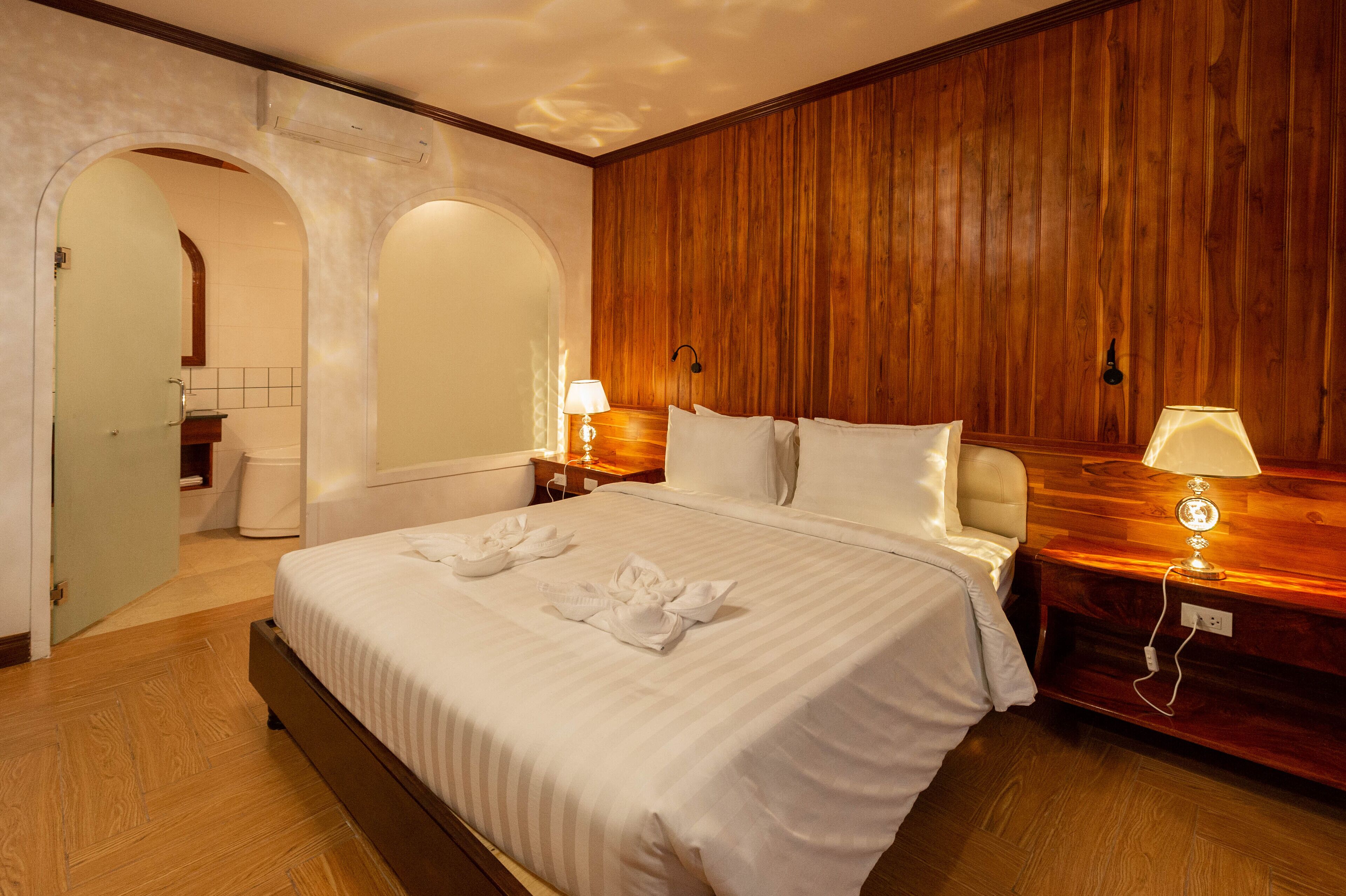 A hotel room with a wooden panel wall, a large bed with white linens, two bedside lamps, and a wooden nightstand.