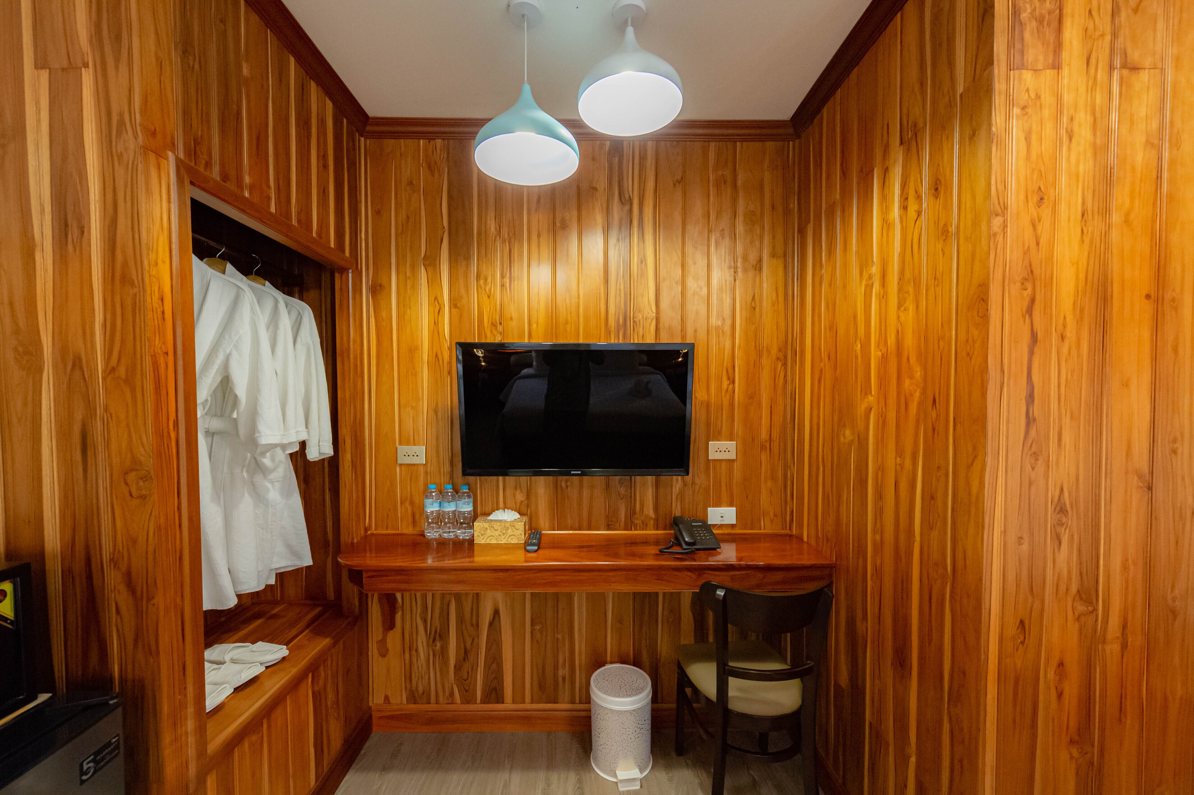 A wooden-paneled room with a TV mounted on the wall, a desk with a phone, a chair, and a shelf with towels and toiletries.