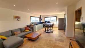 A modern living room with a grey sofa, a wooden coffee table, and a dining area with a table and chairs. There is a kitchen with a window overlooking a scenic view. - Villa Indépendante - Piscine Privée - Clim - Vue Exceptionnelle (Orgnac l'Aven)