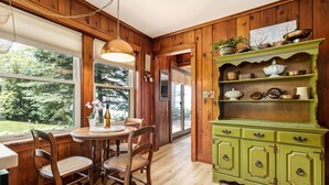 Un intérieur en bois avec une table et des chaises, un buffet vert contenant de la vaisselle, et une vue sur l’extérieur.