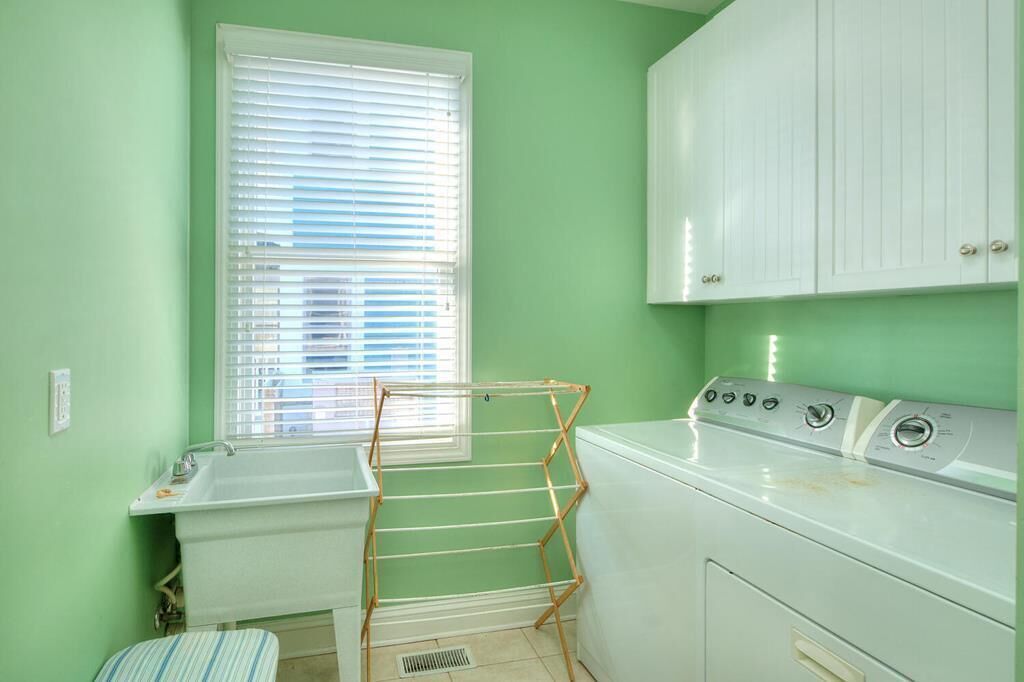 A laundry room with a washing machine, a drying rack, a window with blinds, and a sink.