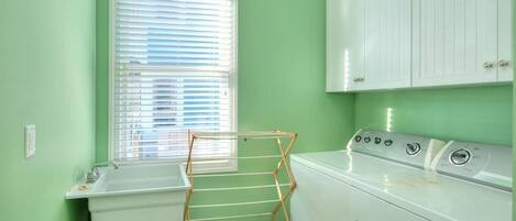 A laundry room with a washing machine, a drying rack, a window with blinds, and a sink.