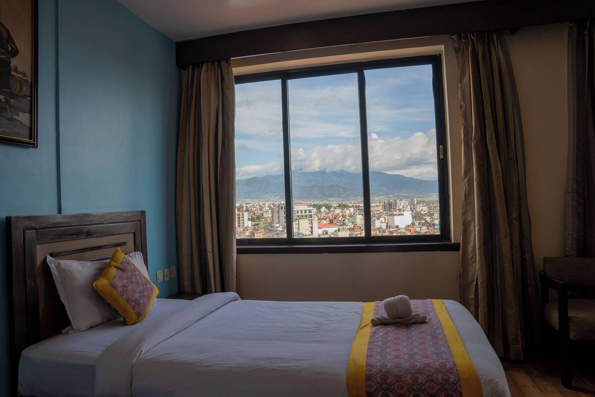A hotel room with a large window, a bed with a patterned bedspread, a chair, and a view of a cityscape.