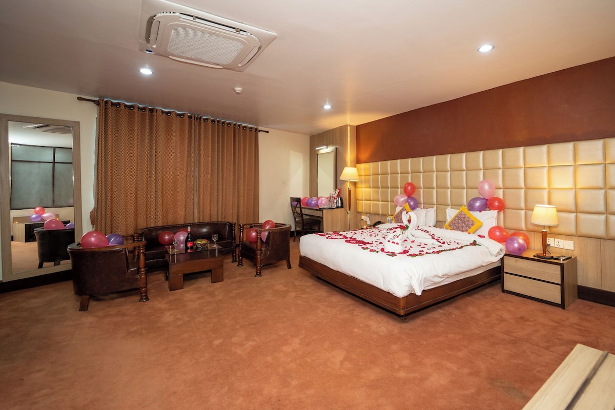 A hotel room with a large bed, a wooden headboard, a bedside table, a nightstand, a chair, a desk, and a mirror.