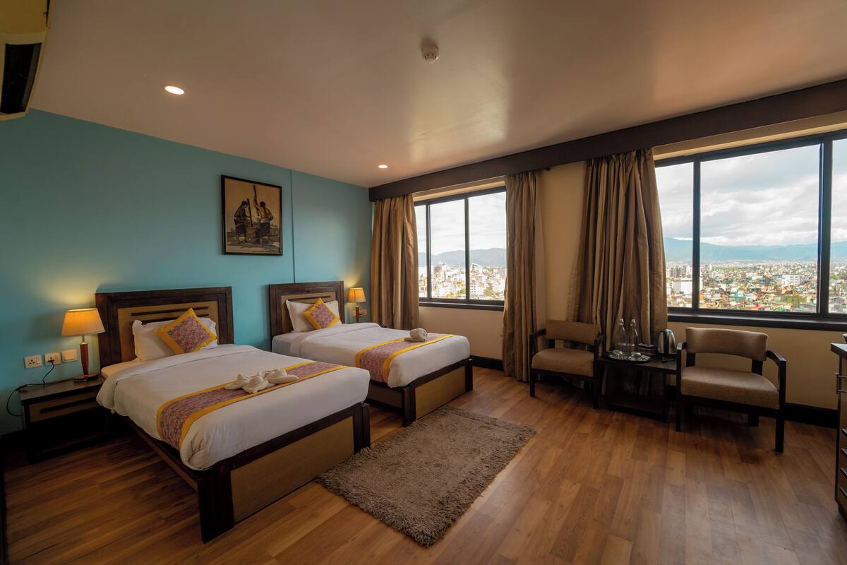 A hotel room with two beds, a desk, a chair, and a large window with a city view.