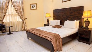 A hotel room with a large bed, two bedside tables with lamps, a small table, and a chair. - Dees Hotels (Lagos)